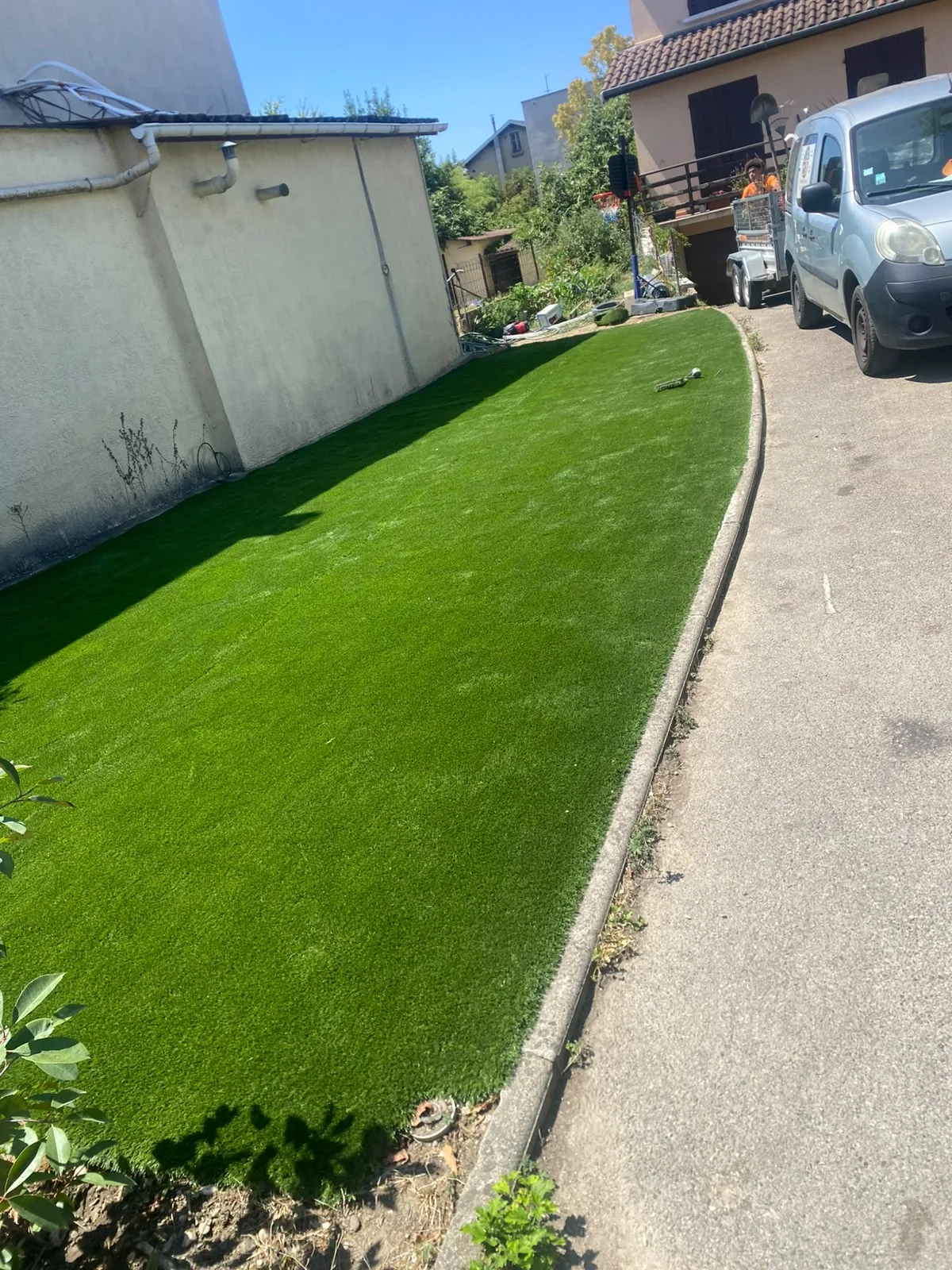 Après — la même bande transformée en pelouse de gazon synthétique posée au pied du mur, jusqu'à la bordure de l'allée